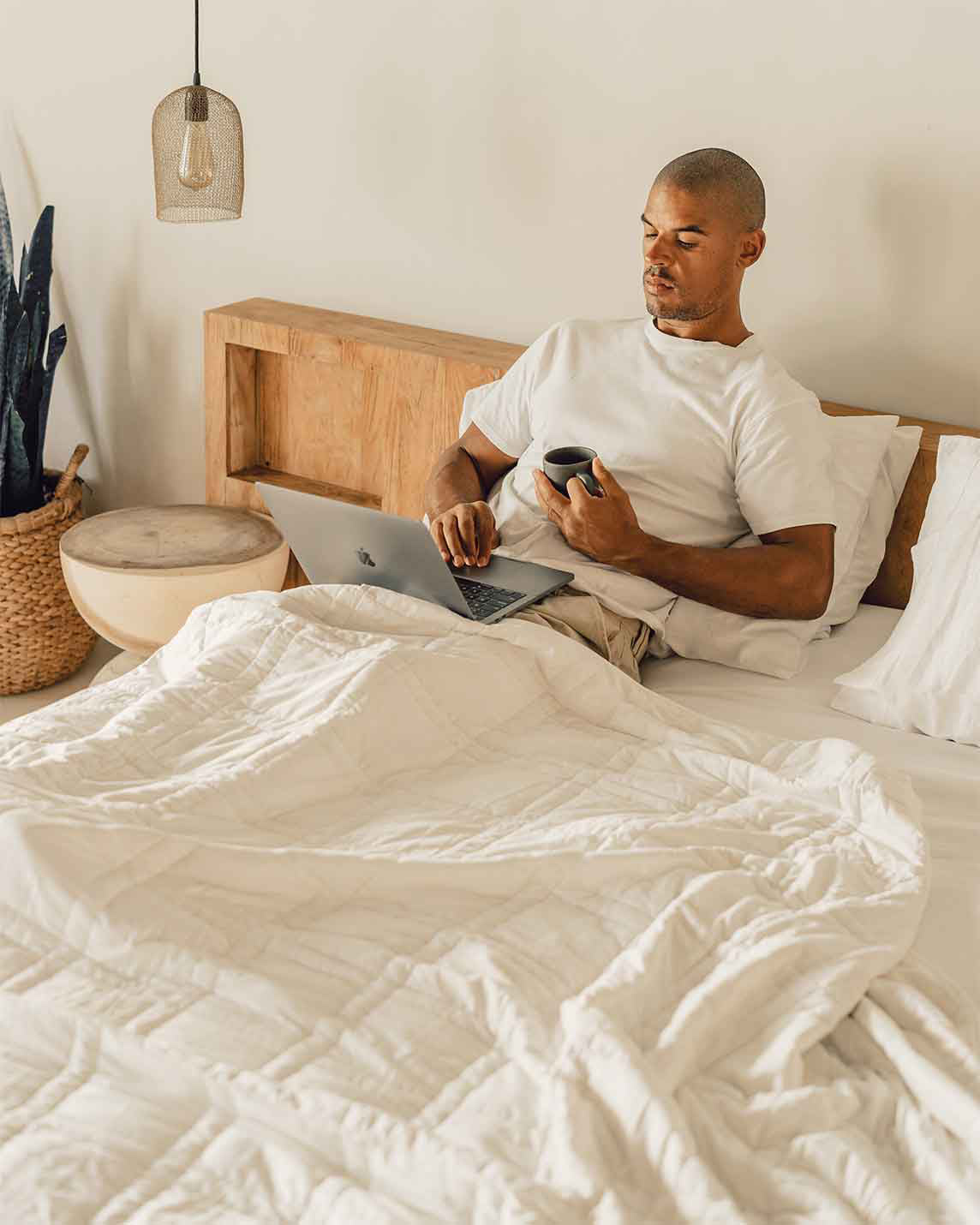Man drinking coffee and using a laptop in bed under the Baloo weighted cotton comforter
