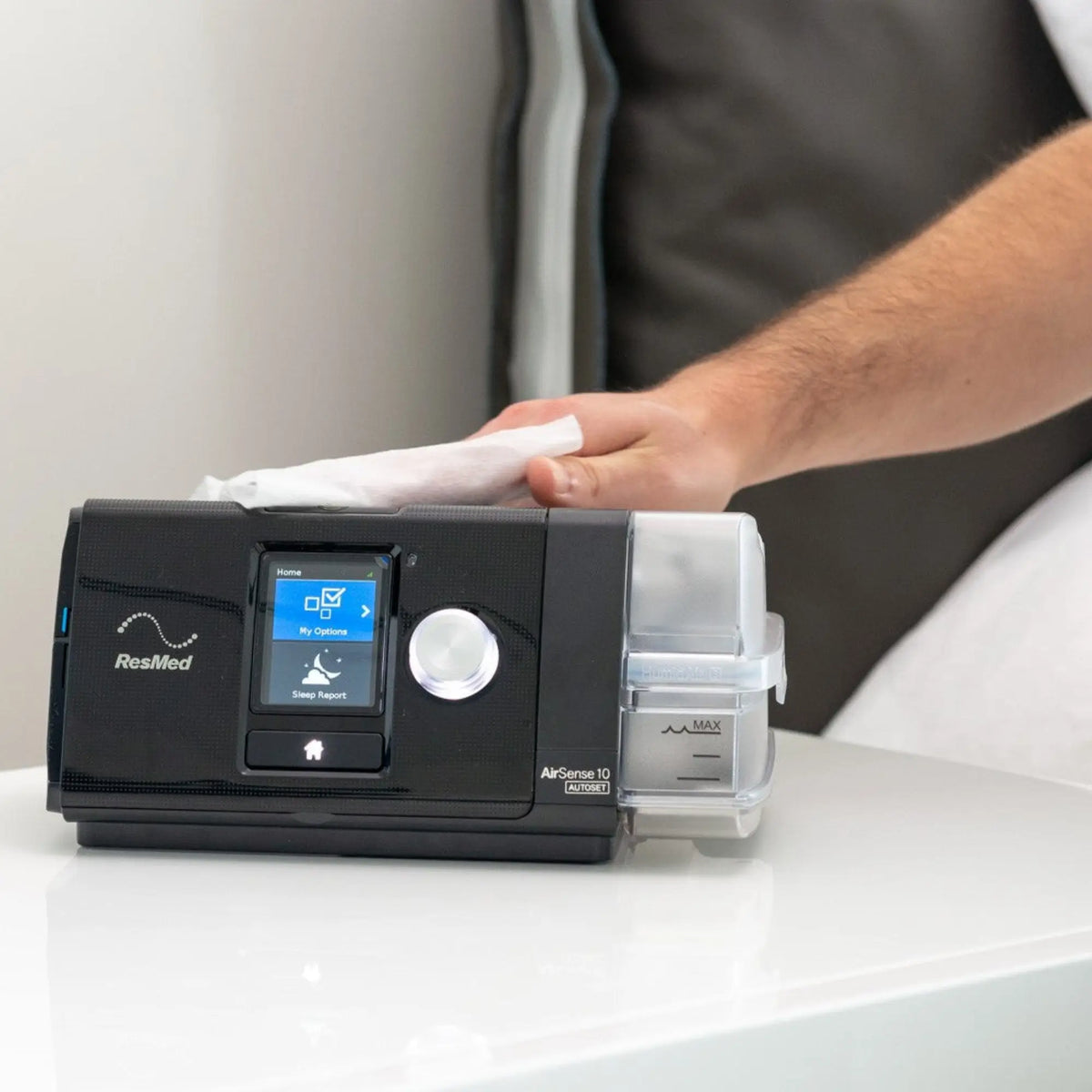 Person wiping ResMed AirSense 10 AutoSet CPAP machine with a white cloth on a bedside table