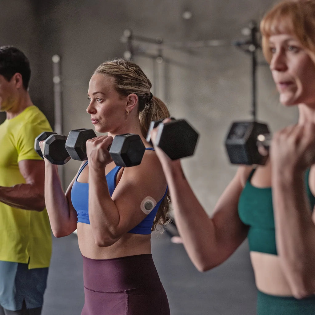 Adults lifting dumbbells at the gym, one wearing a Dexcom Stelo biosensor on her upper arm