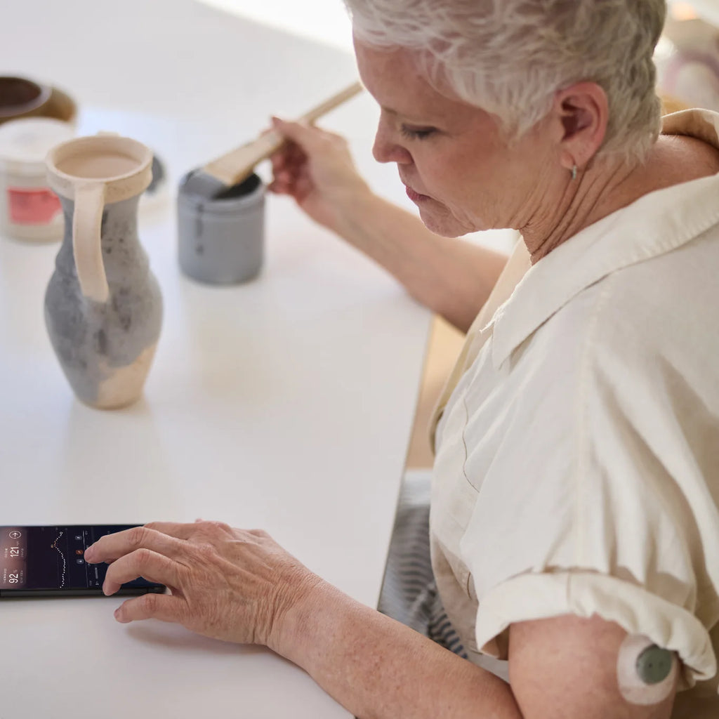 Woman painting ceramics checks the Stelo app on her phone, Dexcom Stelo biosensor on her upper arm