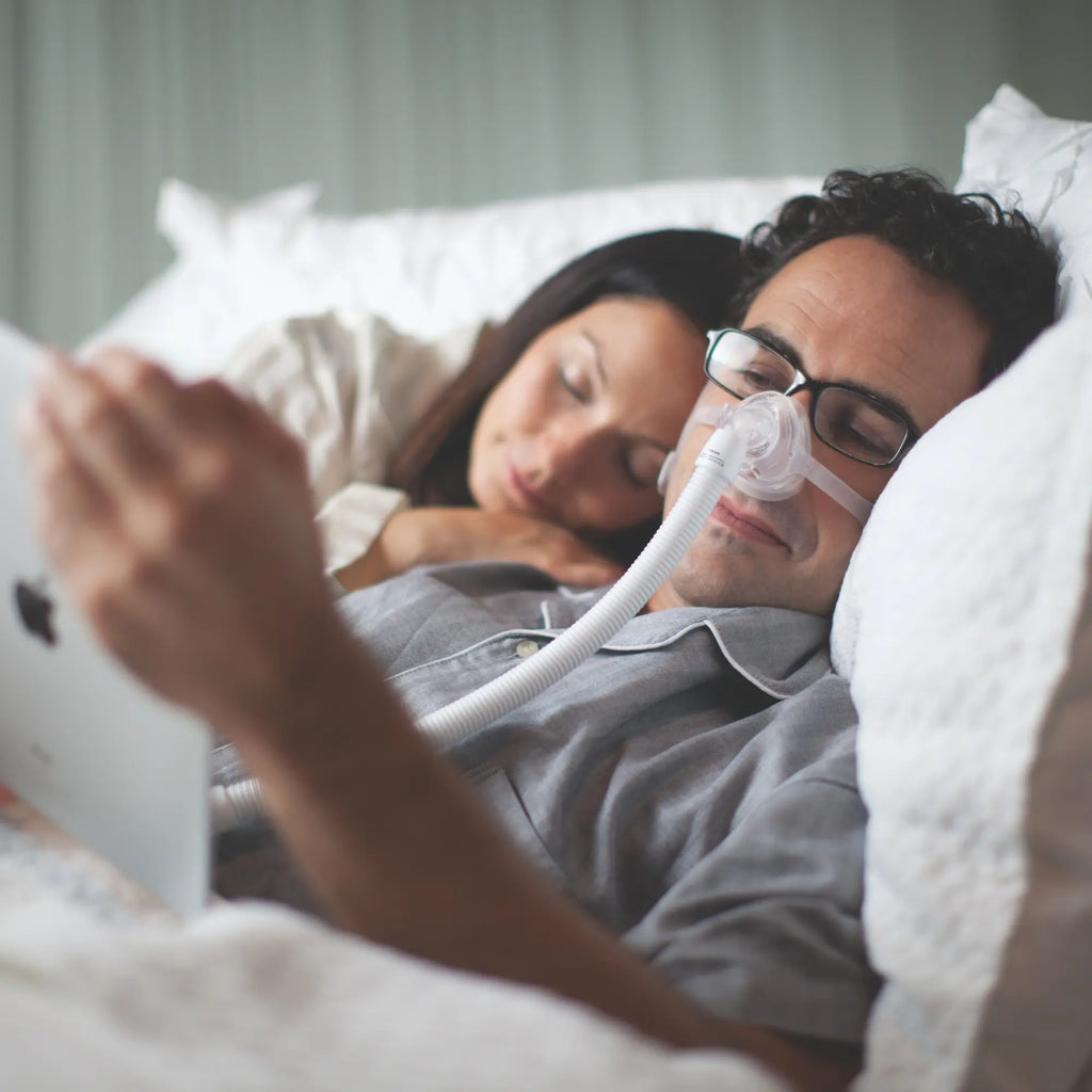 Man wearing Wisp nasal CPAP mask reads a tablet in bed beside his sleeping partner