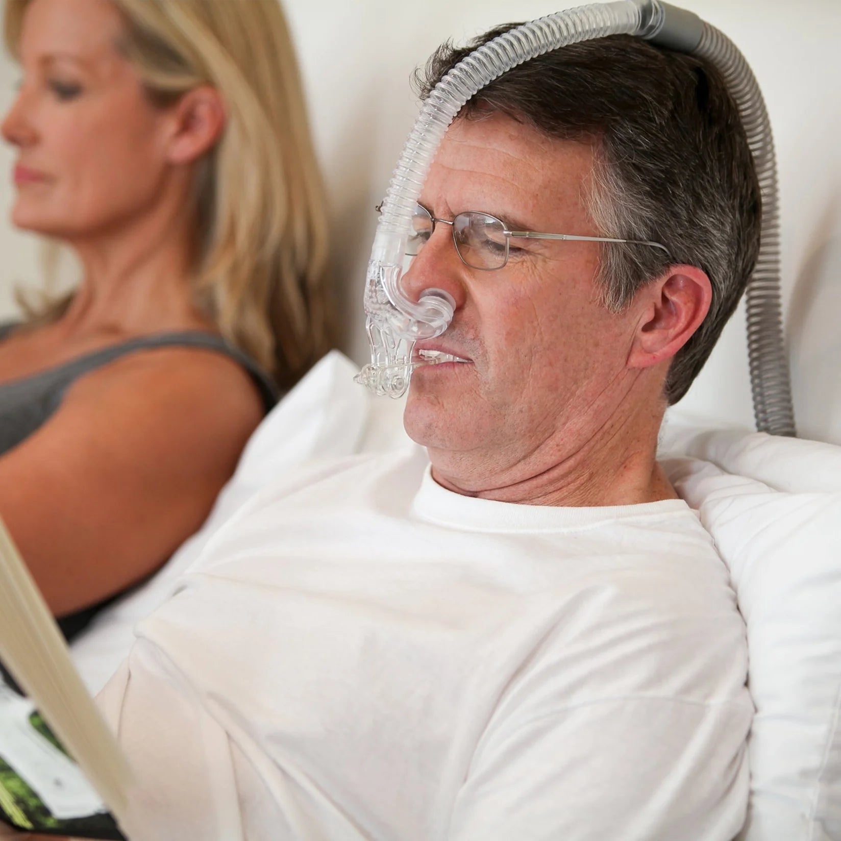 Man with glasses reading in bed wearing myTAP PAP CPAP mask, demonstrating strapless comfort while awake