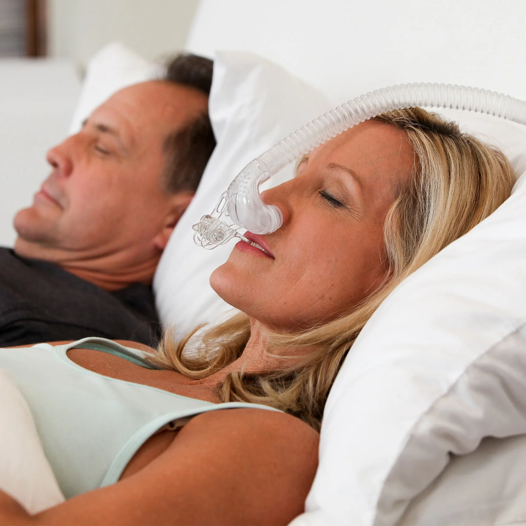 Woman sleeping on her side wearing myTAP PAP strapless nasal pillow mask, partner asleep undisturbed beside her