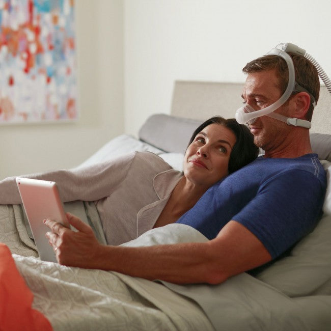 Couple in bed and man using DreamWisp Nasal Mask while reading.